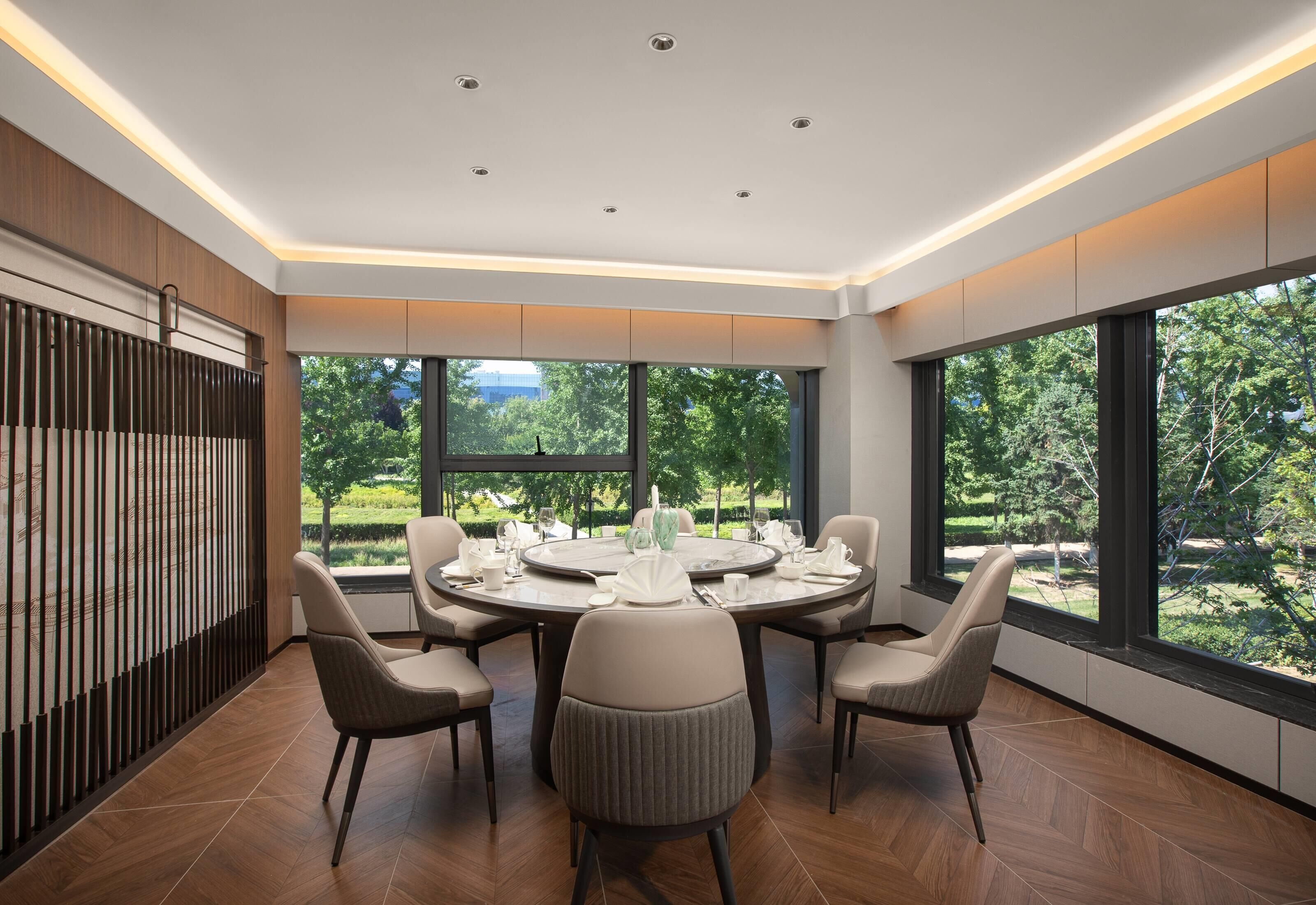 A modern dining area with a round table set for six, surrounded by six chairs. The room has large windows offering a view of greenery outside.