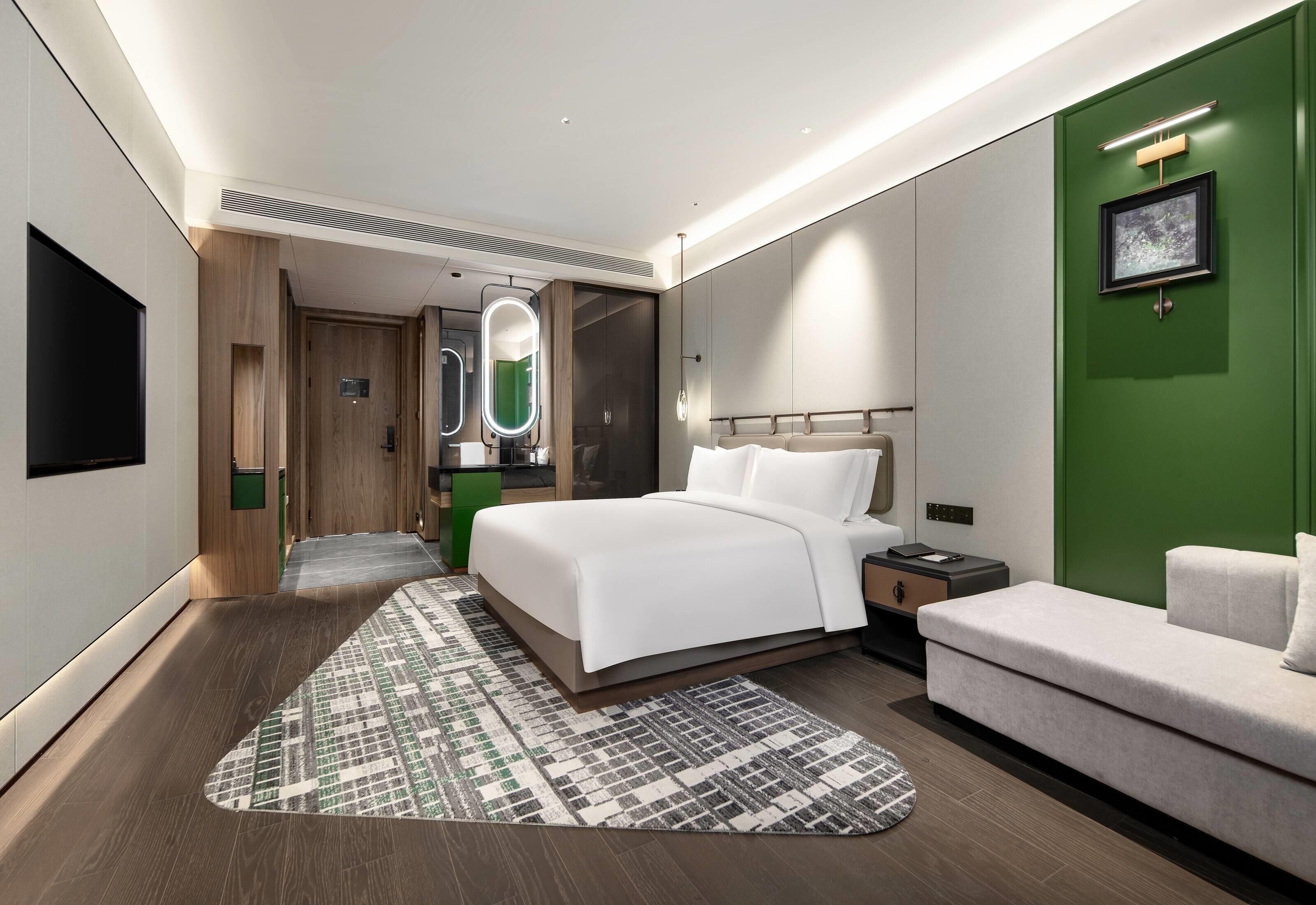 Quarto de hotel moderno com uma cama grande, um sofá, uma parede verde decorativa e um espelho iluminado.