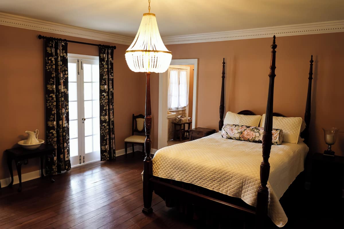 A four-poster bed with a quilted bedspread, a chandelier, and a wooden nightstand with a lamp.