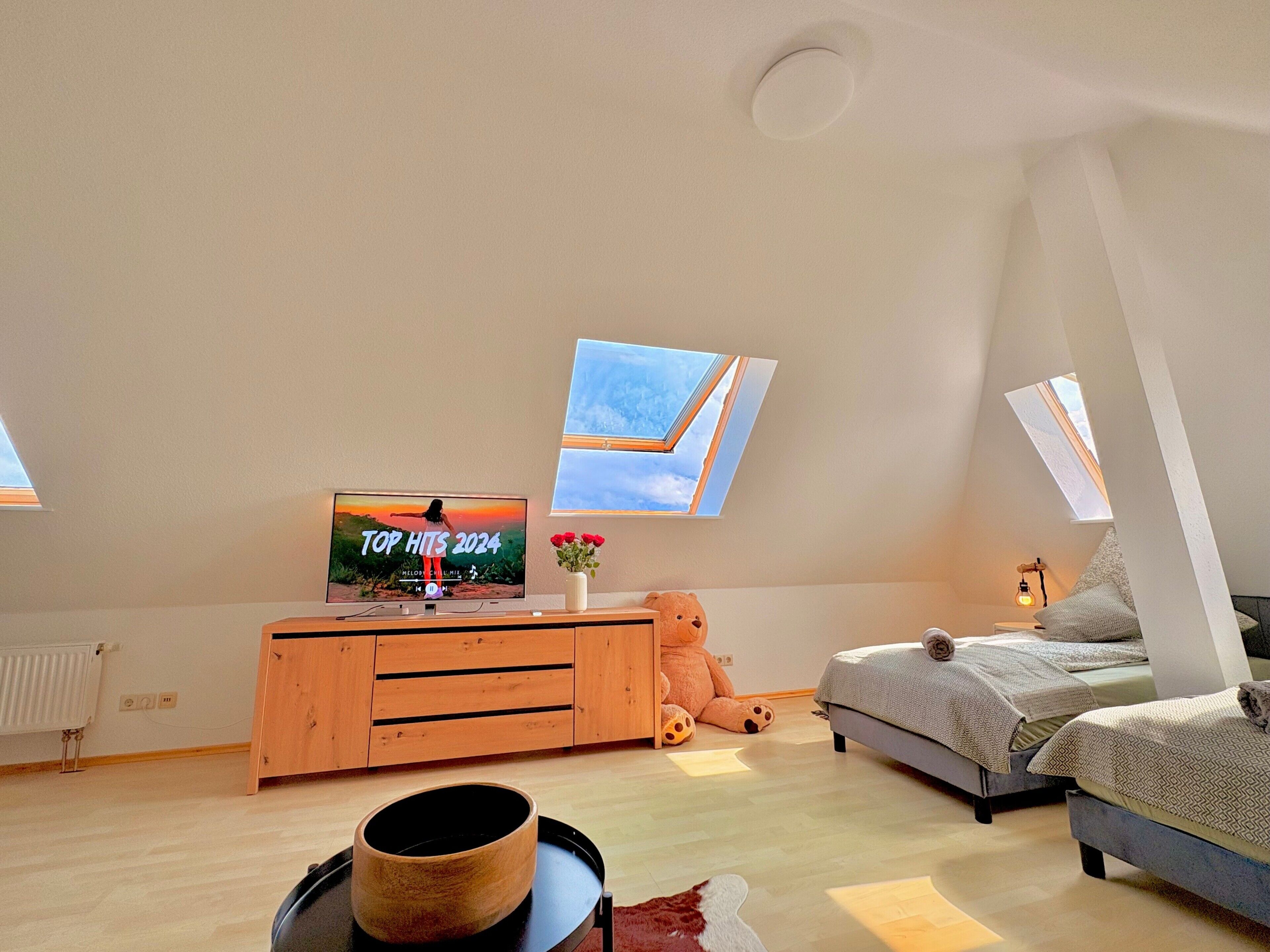 A loft bedroom with a skylight, a television displaying 'Top Hits 2024', a wooden dresser, a teddy bear, and a bed with a pillow.