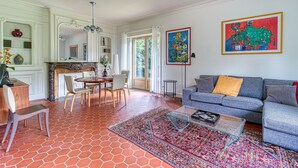 A living room with a sofa, a coffee table, and a patterned rug. There is a dining area with chairs and a table. - Le Logis - Charmante Maison Proche Plage (Téteghem-Coudekerque-Village)