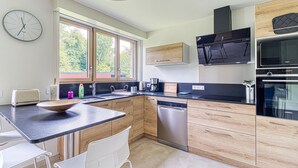 A modern kitchen with wooden cabinets, a dishwasher, a built-in oven, and a large window providing natural light. - Le Logis - Charmante Maison Proche Plage (Téteghem-Coudekerque-Village)