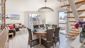 A modern living room with a dining area, featuring a staircase, wicker furniture, and large windows. - L'appart du Val - 100 m de la plage (Pléneuf-Val-André)