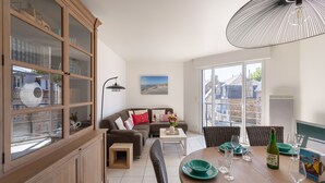 A modern living room with a dining area, featuring a wooden cabinet, a sofa, a dining table with chairs, and a large window offering a view of the outside. - L'appart du Val - 100 m de la plage (Pléneuf-Val-André)