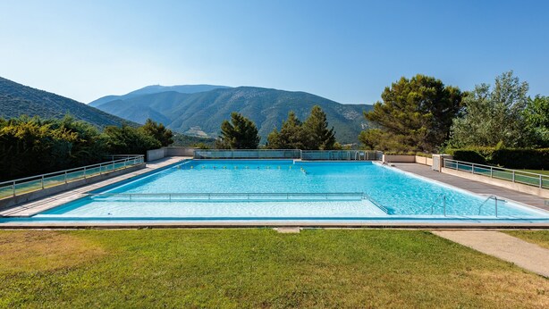 A rectangular swimming pool with a clear blue water surface, surrounded by a grassy area and trees, with mountains in the background.