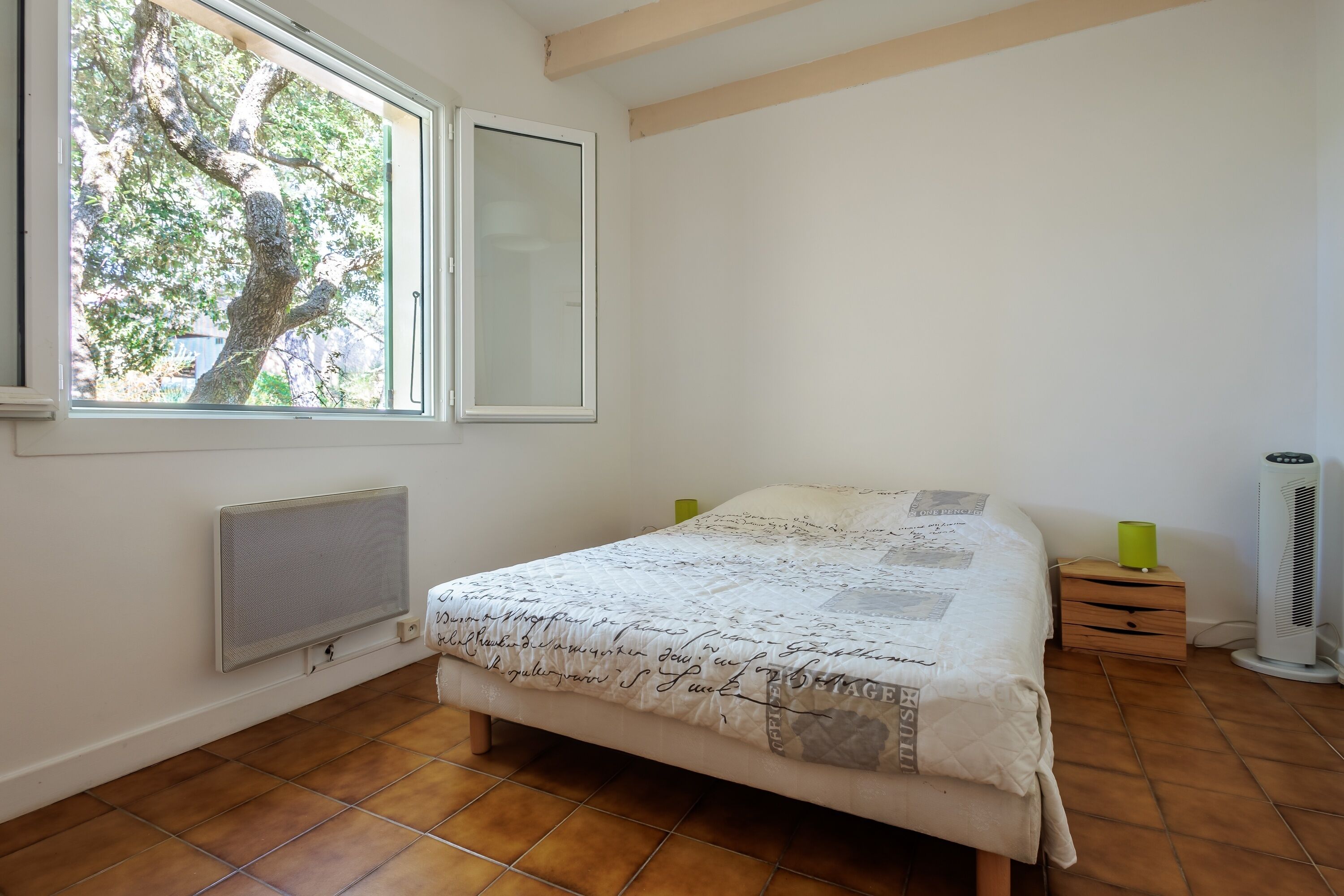 A bedroom with a bed, a radiator, a window, a nightstand, and a standing heater.