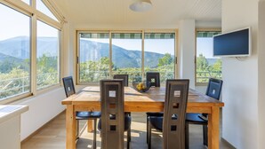 A dining area with a wooden table and chairs, large windows with a mountain view, and a flat-screen TV mounted on the wall.