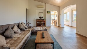 A spacious living room with a wooden ceiling, a sofa with cushions, a coffee table, and a kitchen area in the background.