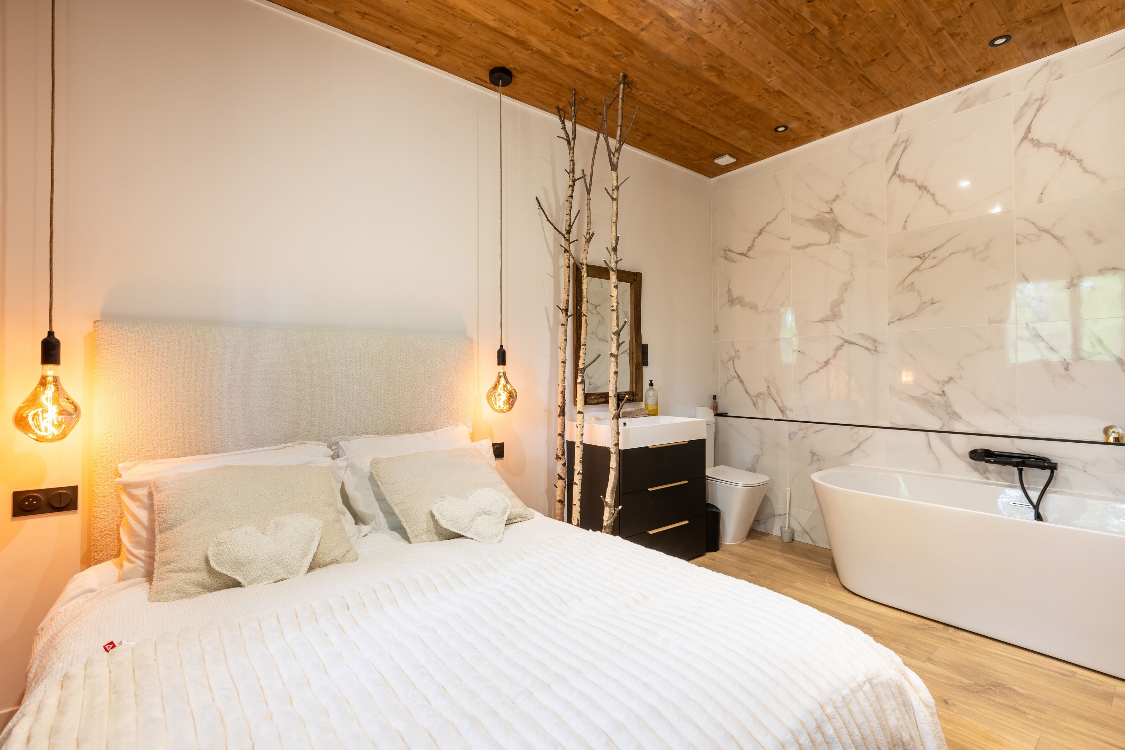 Ein modernes Schlafzimmer mit einem großen Bett, ein weißes Badezimmer mit freistehender Badewanne und einer Holzbalkendecke.