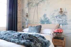 Un dormitorio con cama, mesita de noche, un jarrón con flores y un mural en la pared.