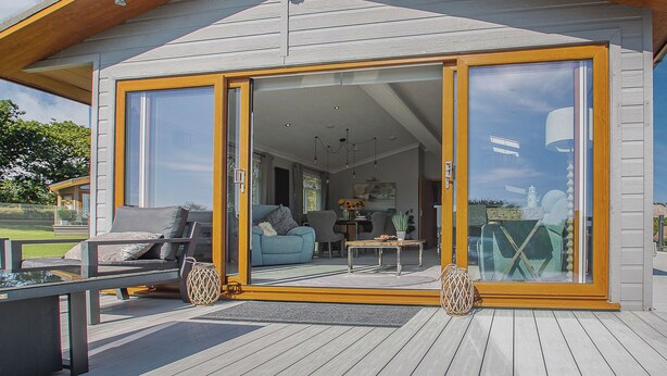 Una casa moderna con un amplio acceso por puerta de vidrio corredera, una terraza con mobiliario exterior y un interior visible que incluye un sofá y una mesa.