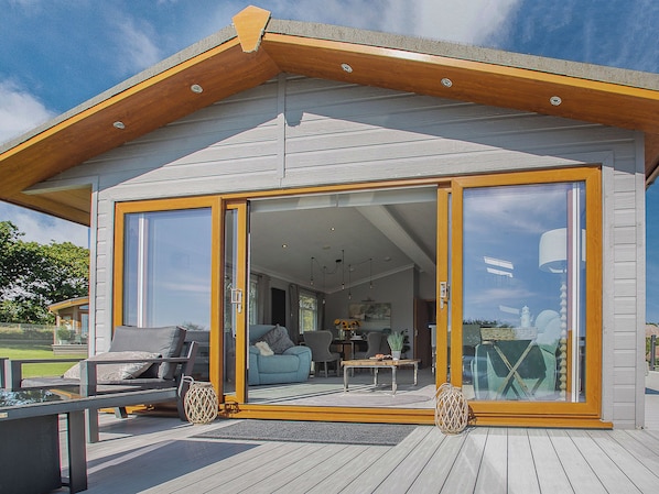 A modern house with a large sliding glass door entrance, a deck with outdoor furniture, and a visible interior with a sofa and table.