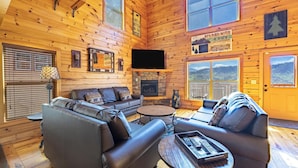 A cozy log cabin interior with a fireplace, leather sofas, and a large window offering a scenic view.