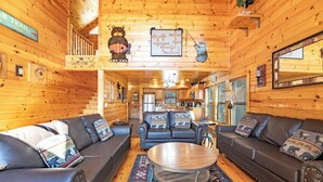 A log cabin interior with a central seating area featuring two sofas and a coffee table, wooden walls, a kitchen in the background, and a sign indicating a 2190-mile trail.