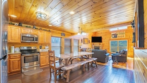 A log cabin interior with wooden floors, a kitchen with stainless steel appliances, a dining area with a wooden table and benches, a living room with a sofa, and a fireplace.