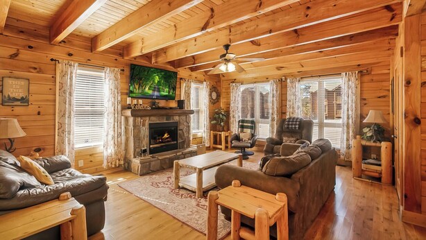 A cozy cabin interior with a stone fireplace, wooden furniture, and a television mounted on the wall. - Center of Attention (Sevierville)
