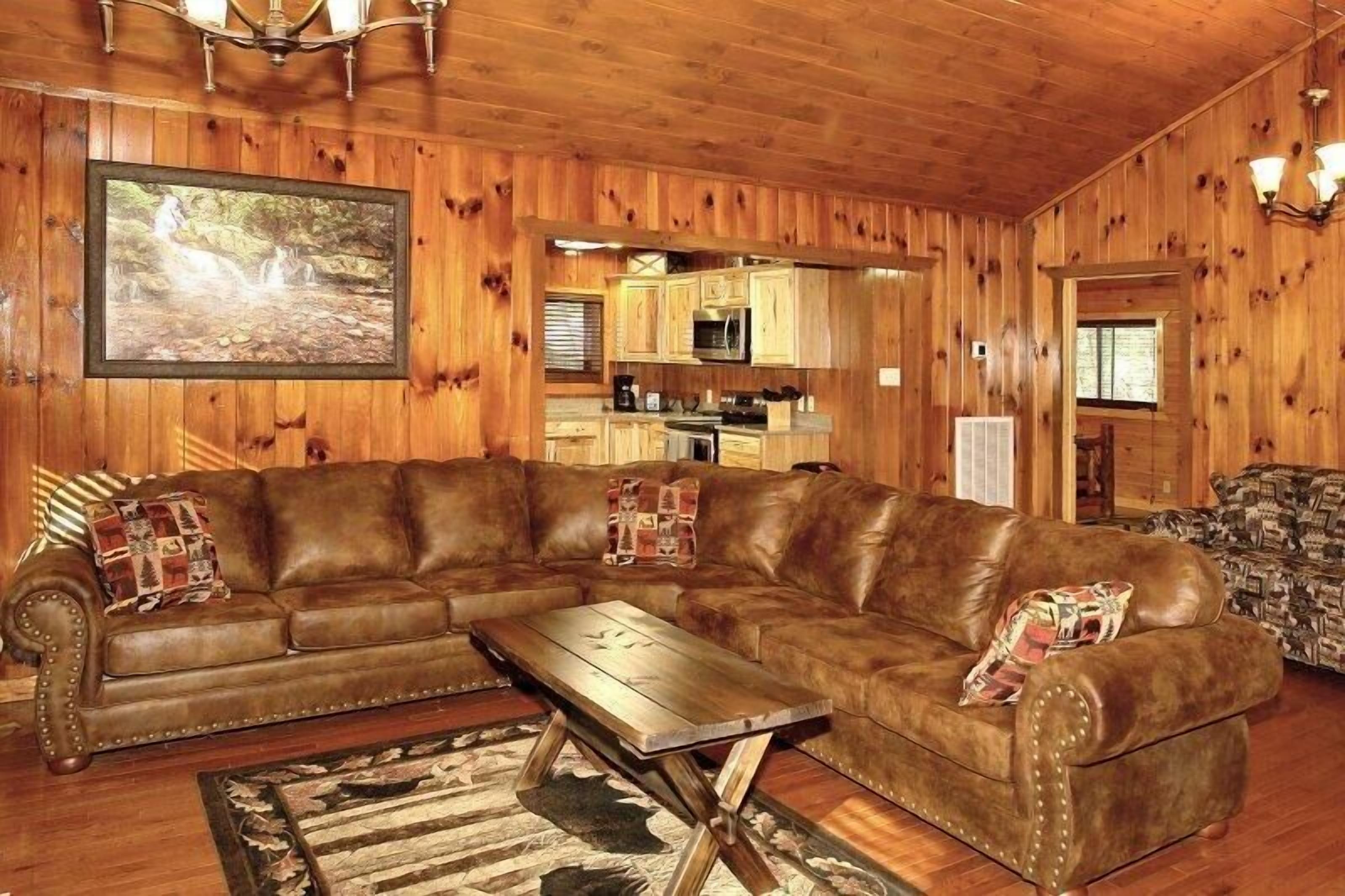 A wooden interior with a leather sectional sofa, a wooden coffee table, and a kitchen area visible through an open doorway.