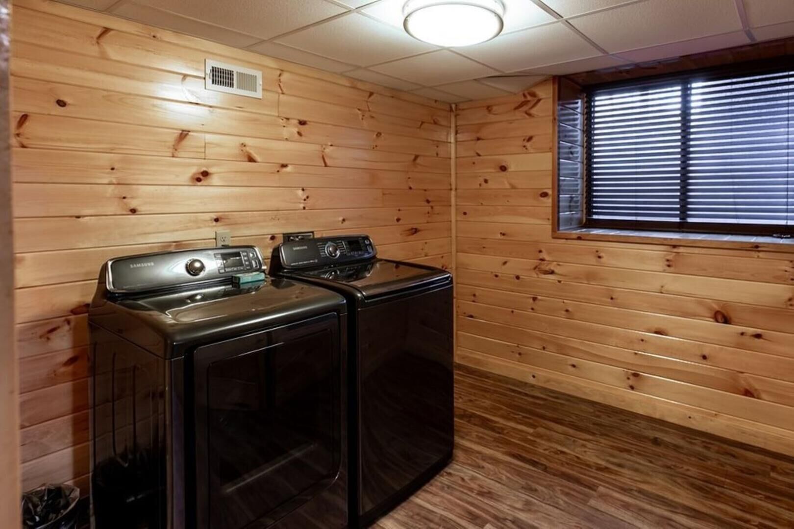 A laundry room with two washers and dryers, wooden walls, a window with blinds, and a ceiling vent.