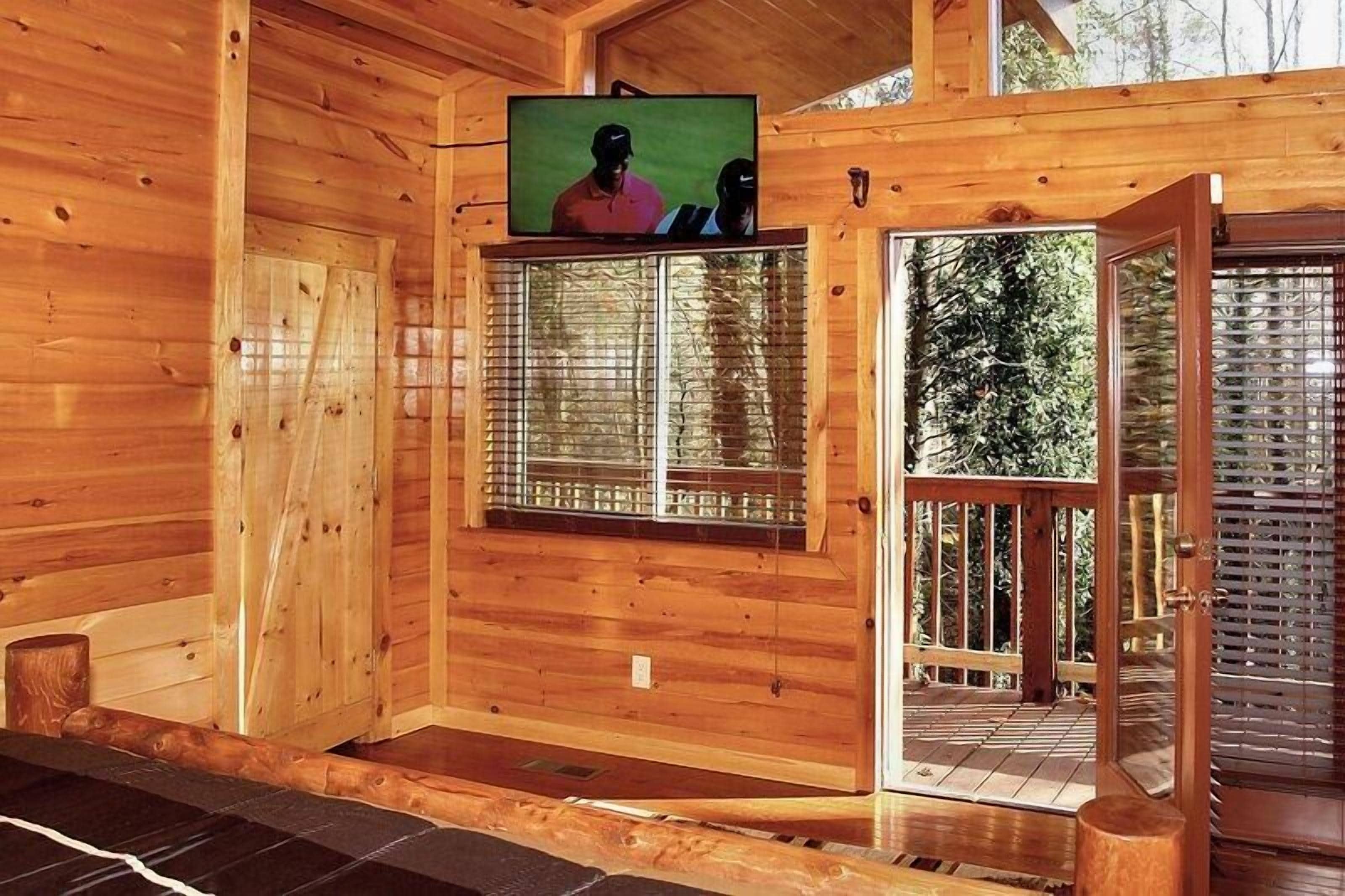 A wooden cabin interior with a television mounted on the wall, a window with blinds, and a door leading outside.