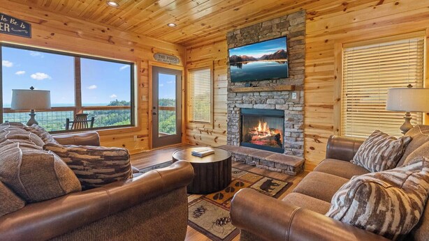 A cozy cabin interior with leather sofas, a stone fireplace, a television mounted above it, and large windows offering scenic views. - Dolly Valley View (Sevierville)