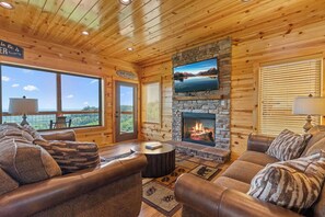 A cozy cabin interior with leather sofas, a stone fireplace, a television mounted above it, and large windows offering scenic views. - Dolly Valley View (Sevierville)