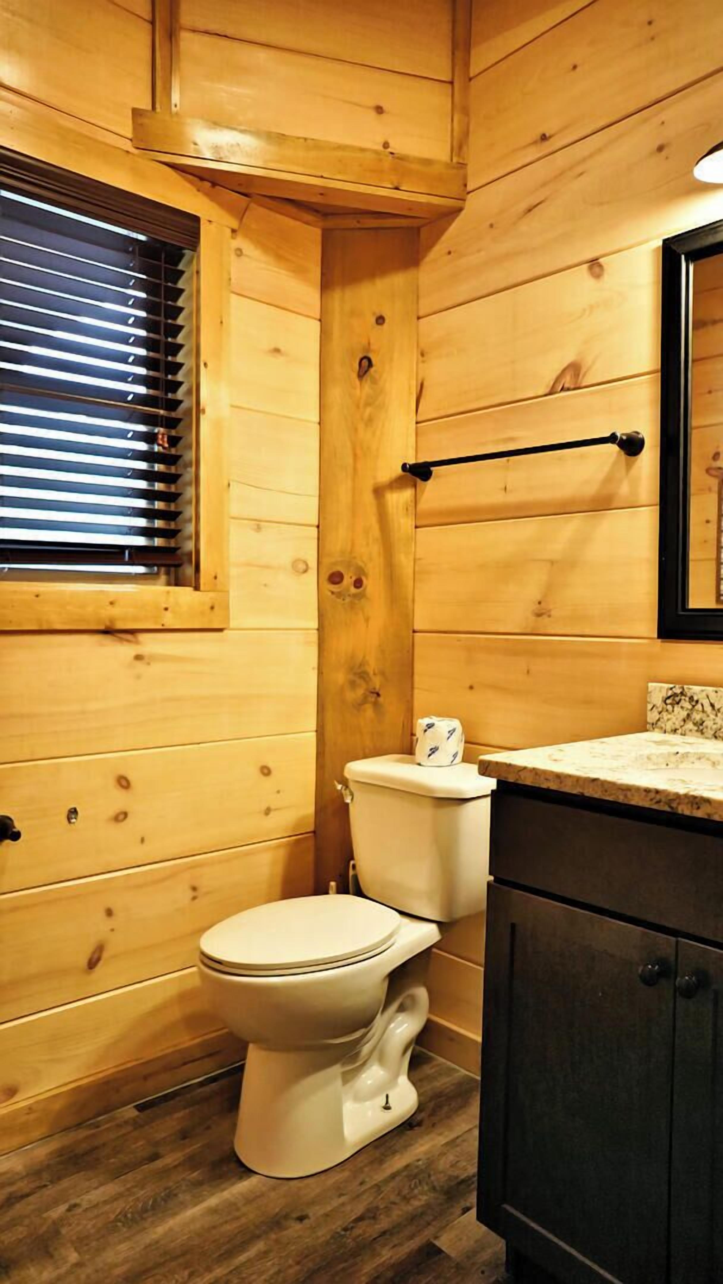 A wooden cabin-style bathroom with a toilet, sink, and window with blinds.