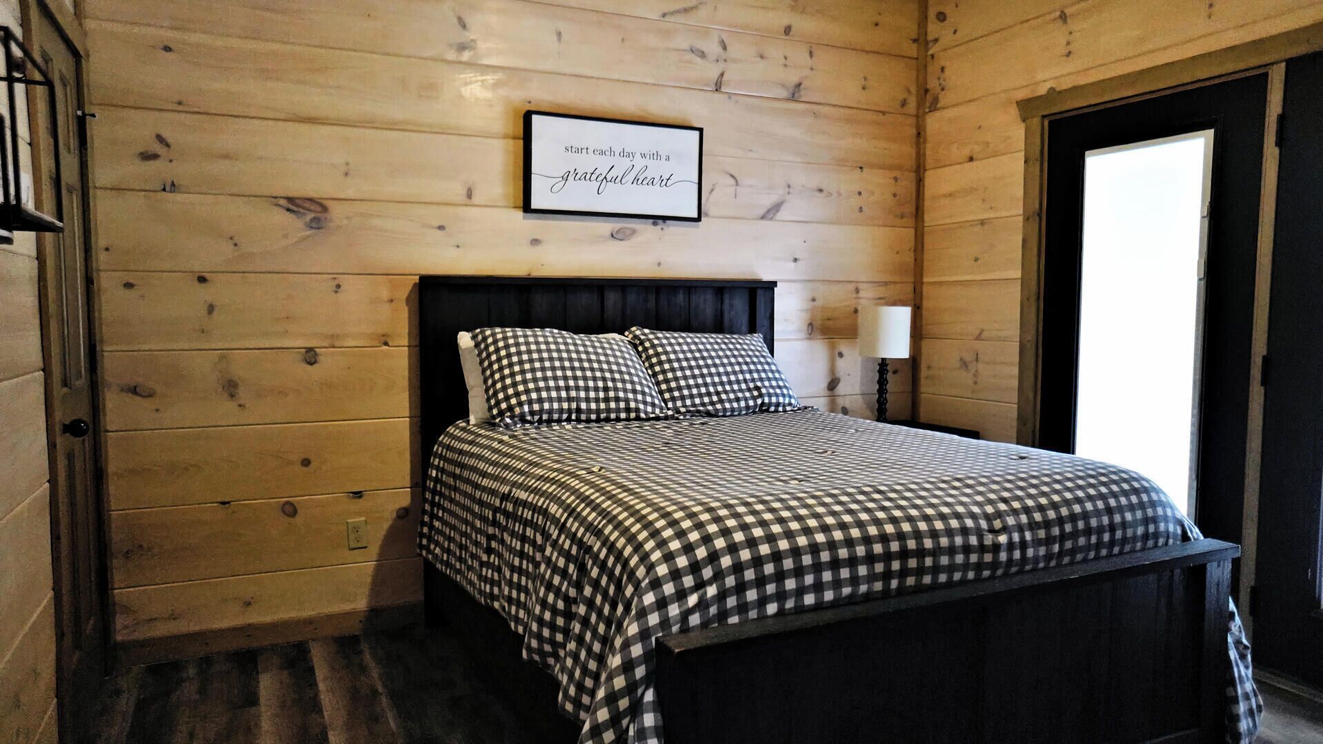 A wooden-paneled room with a bed featuring a checkered bedspread and pillows, a framed inspirational quote on the wall, and a nightstand with a lamp.
