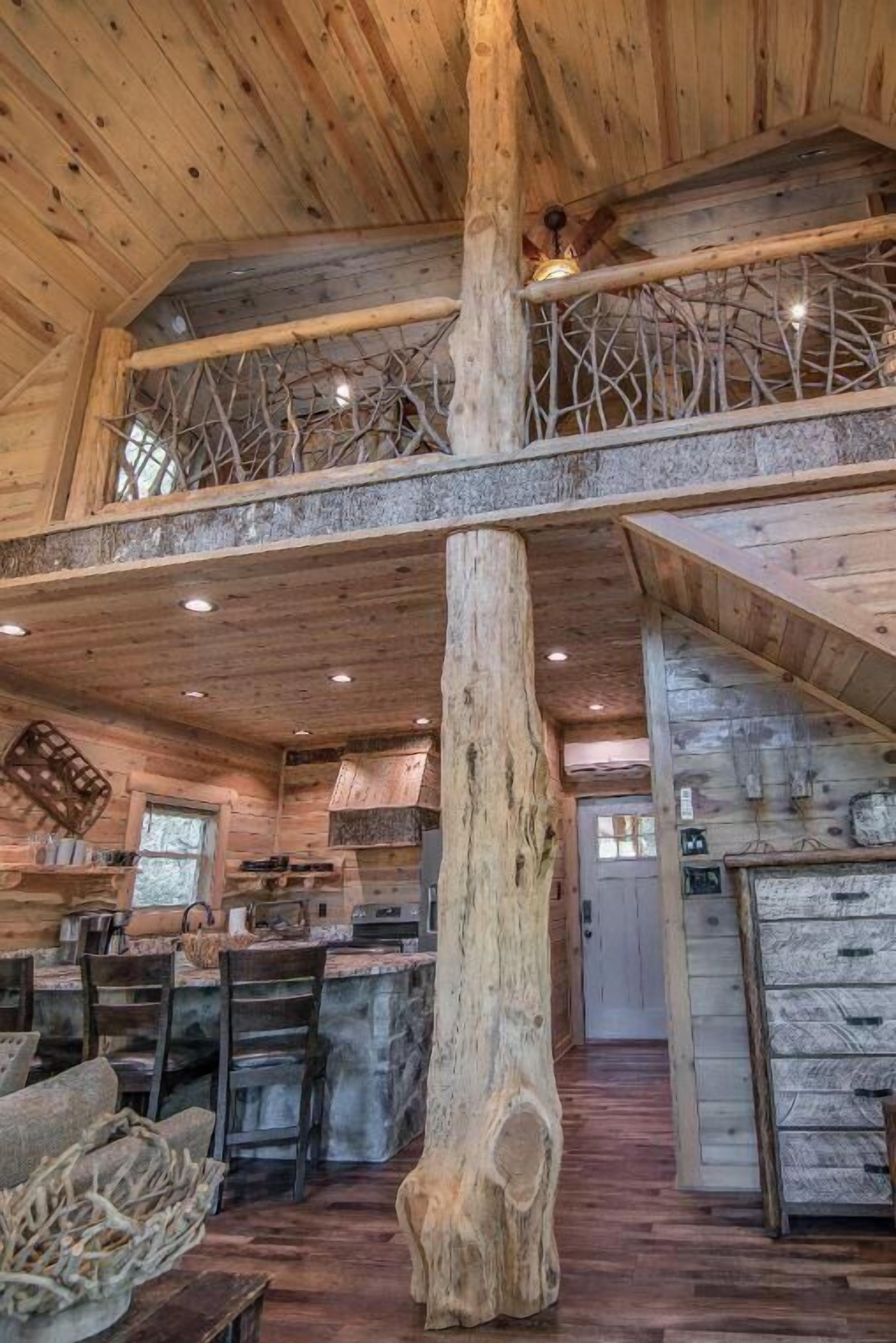 Interior rústico de una cabaña con una viga de madera destacada, una zona de comedor con mesa de piedra y una cocina con armarios de madera.