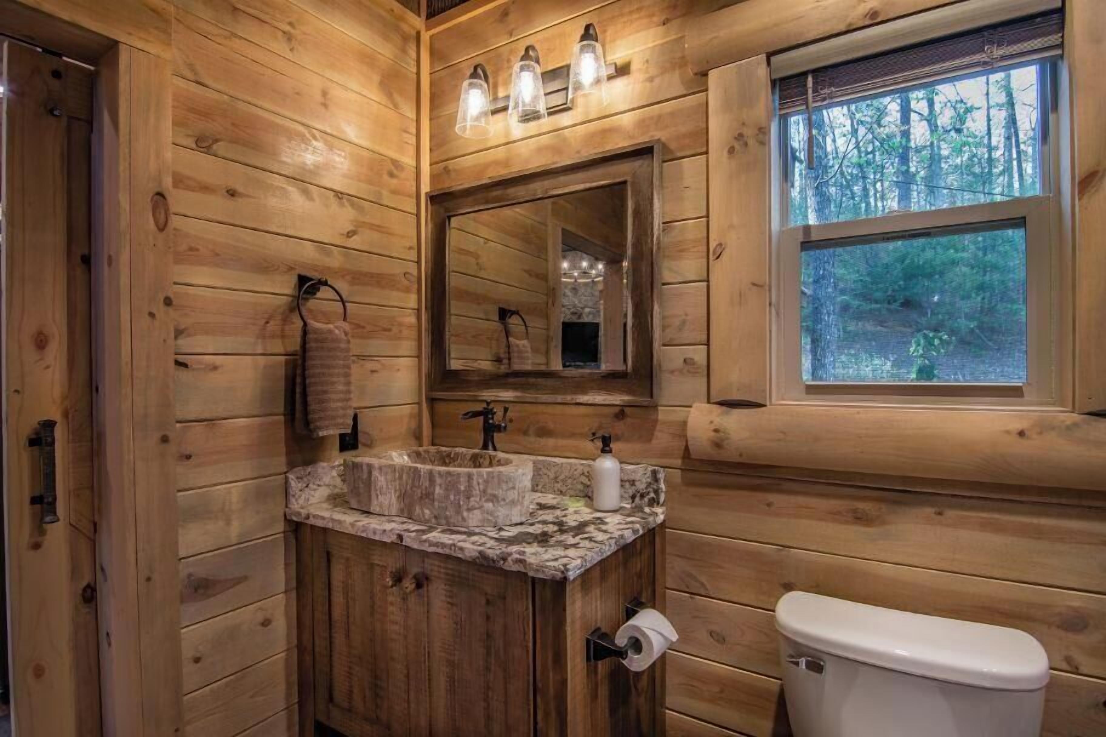 A wooden cabin bathroom with a wooden vanity, a sink, a toilet, and a window with a view of trees.