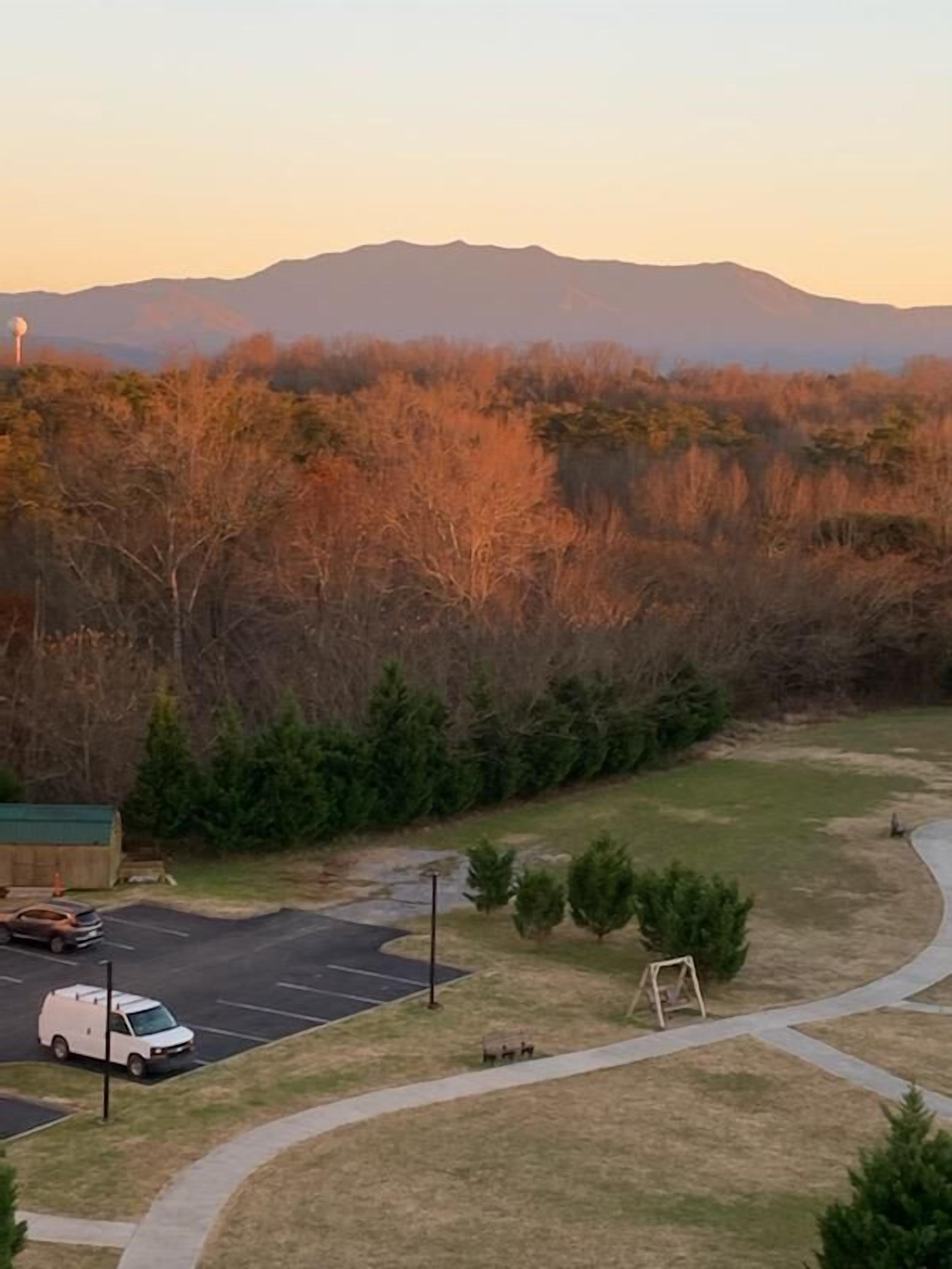 A parking lot with a white van, a swing set, and a view of a forested area with mountains in the background.