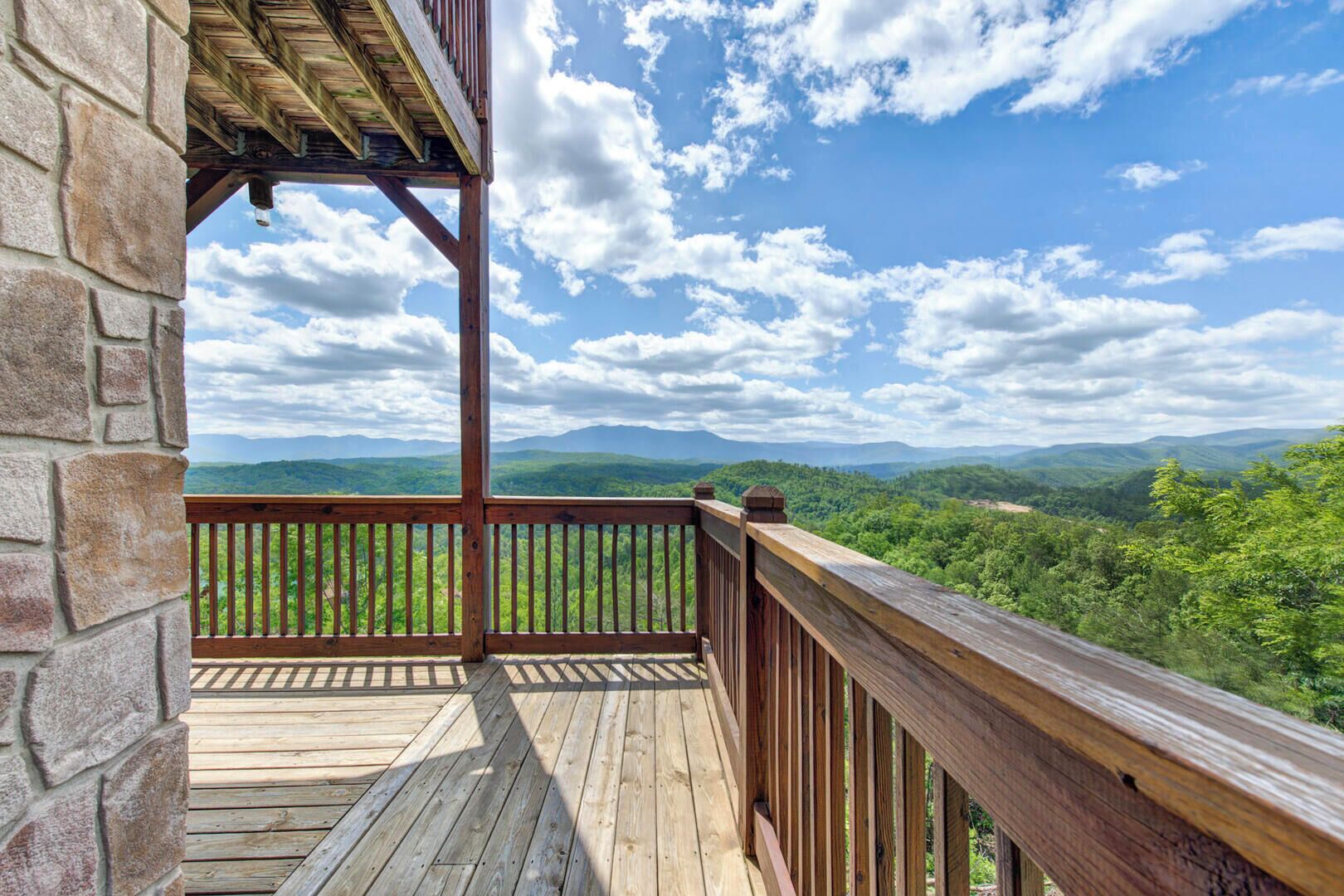 Una terraza de madera con un muro de piedra, con vistas a un paisaje verde exuberante y montañas distantes bajo un cielo parcialmente nublado.