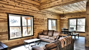 A cozy cabin interior with wooden walls, large windows, a brown leather sofa, a dining table, and a small table with a bicycle wheel base. - Wagon Wheel (Sevierville)