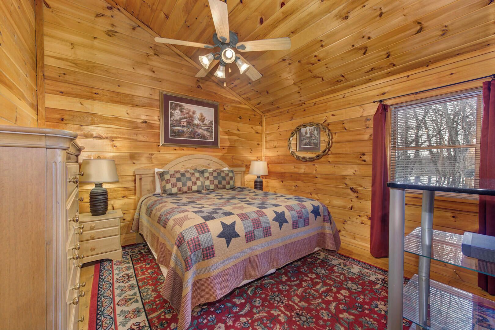 Una habitación de cabaña acogedora, con techo de madera, una cama con colcha, un aparador, una lámpara y una ventana con cortinas.
