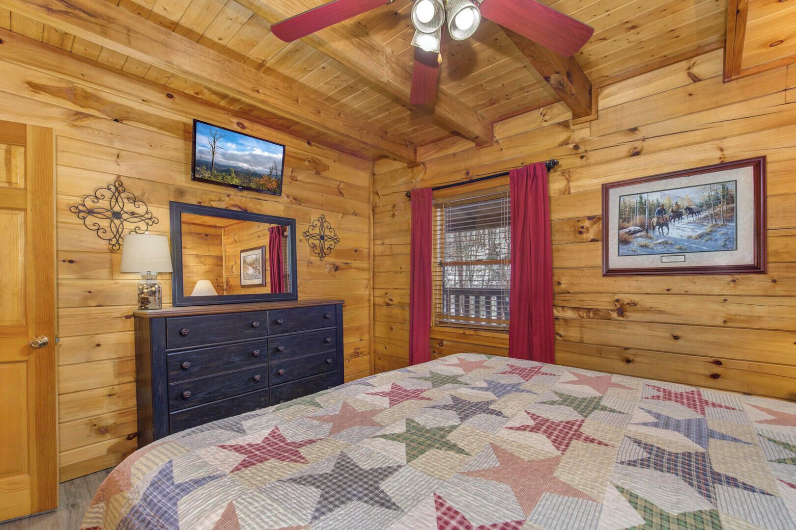 A cozy cabin room with a wooden interior, a bed with a star-patterned quilt, a dresser, a mirror, a television, and a framed picture on the wall.