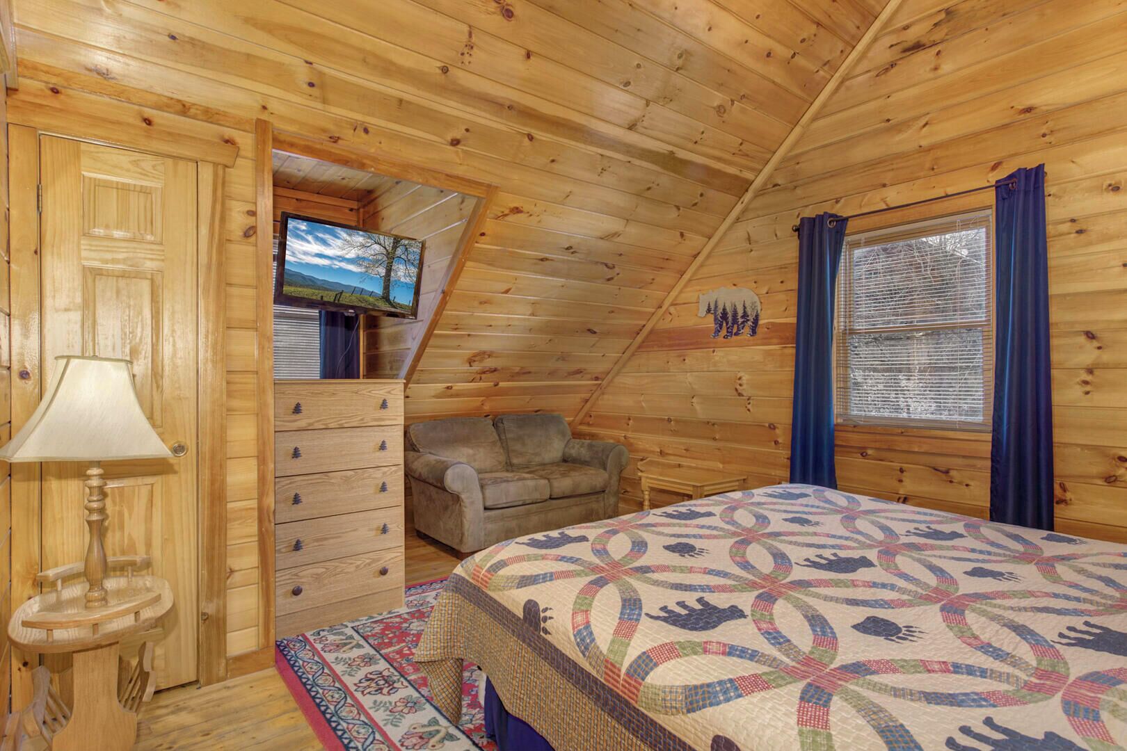 A cozy cabin bedroom with a wooden ceiling, a bed with a patterned quilt, a dresser, a chair, and a television on a wall.