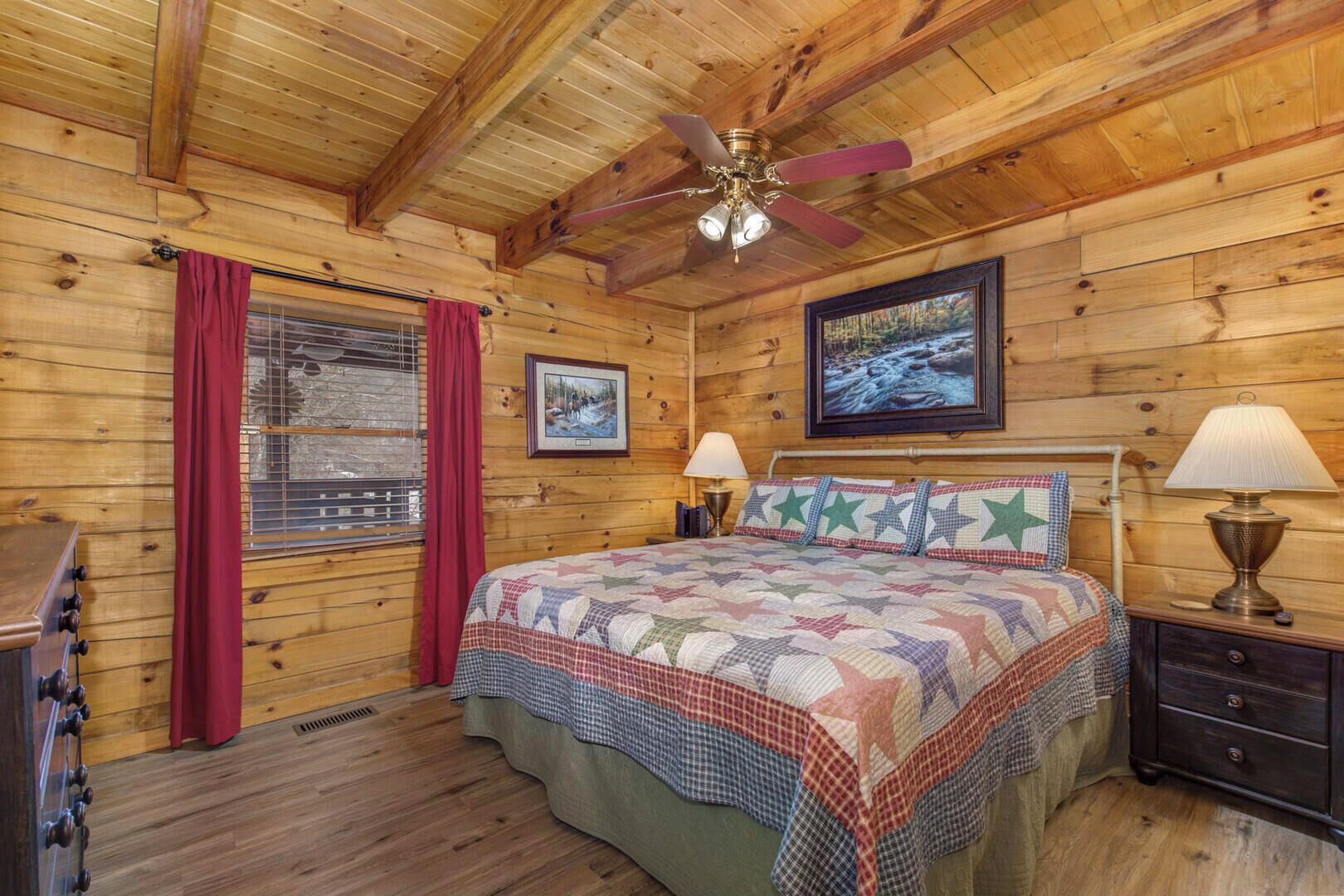 A cozy cabin bedroom with a wooden ceiling, a bed with a quilt, two bedside lamps, and a window with curtains.