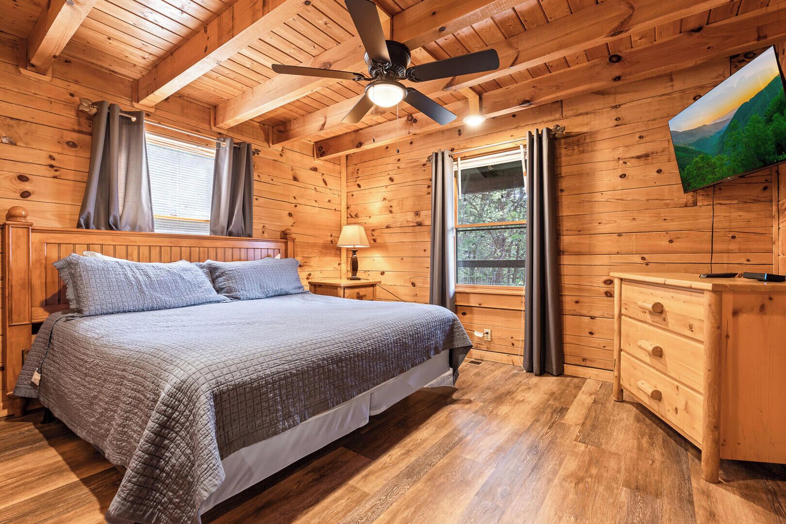 Ein Schlafzimmer einer Holzhütte mit Bett, Nachttisch, Kommode, Deckenventilator und einem Fenster mit Vorhängen.