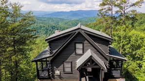 Une cabane en rondins surmontée d’un toit à pignon abrupt, entourée d’une forêt dense et avec des montagnes en arrière-plan.