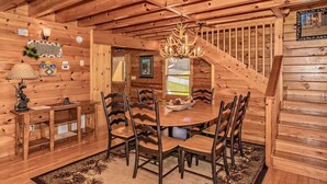 A wooden interior with a round dining table surrounded by chairs, a chandelier, and a staircase. - A Mountain Dream (Sevierville)