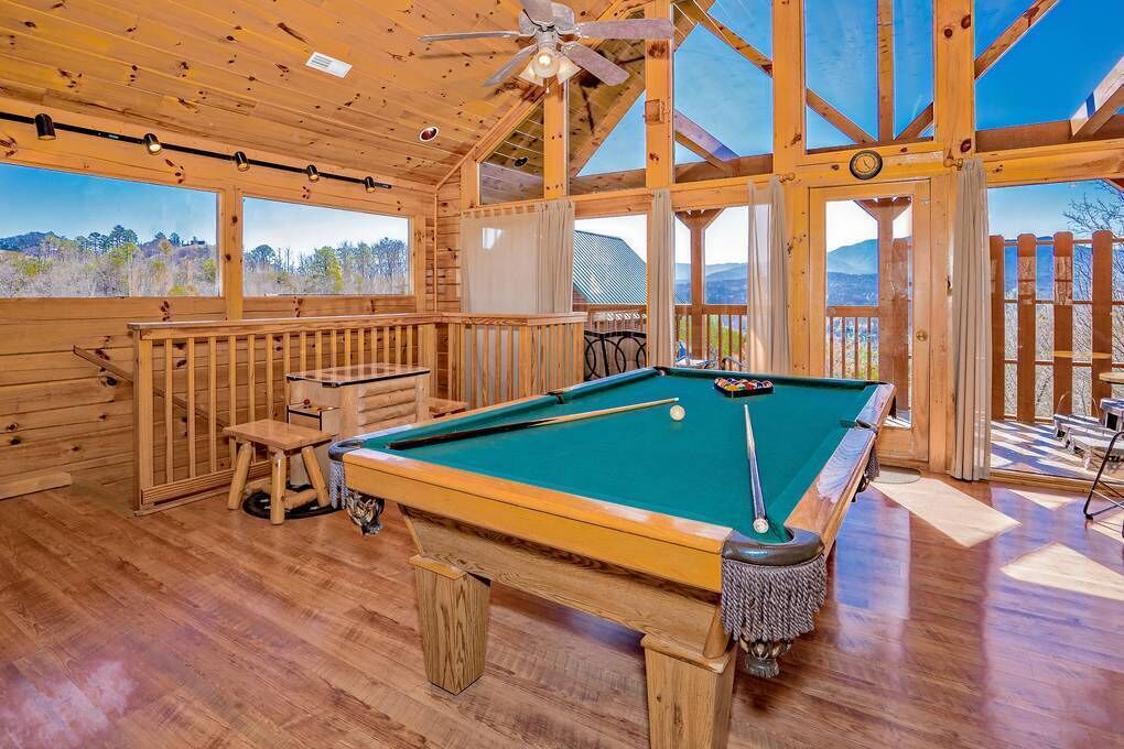 A wooden interior with a pool table, a ceiling fan, and large windows offering a view of the outdoors.