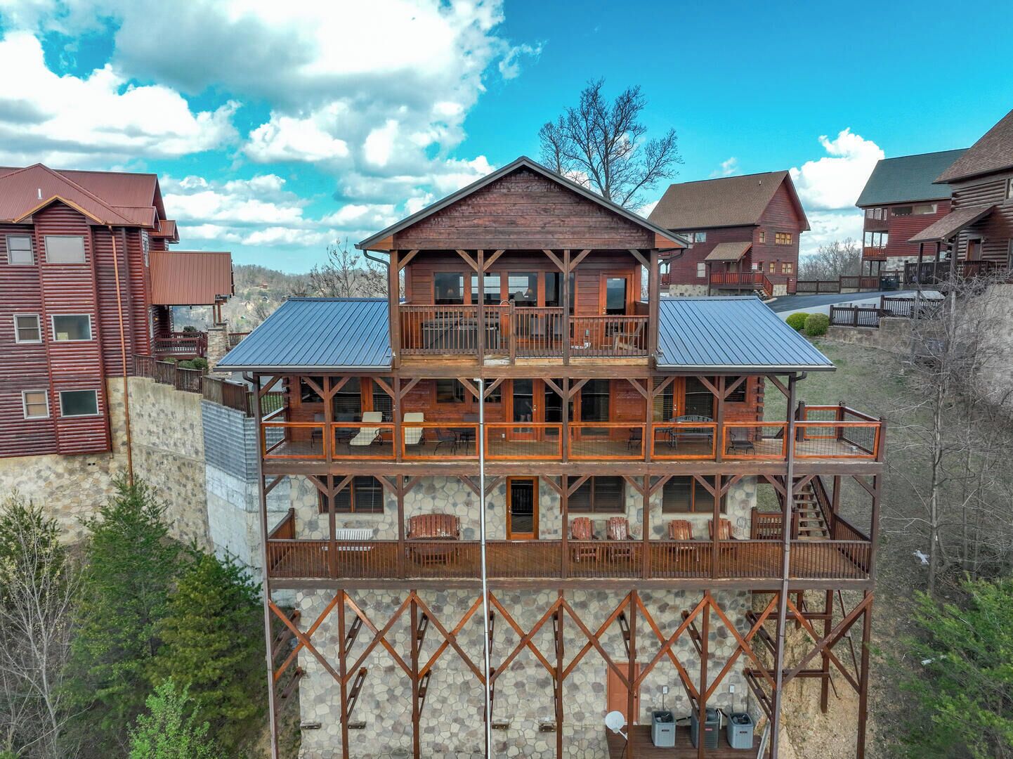 A multi-story log cabin with a balcony on the upper floor, stone foundation, and a mountainous backdrop.