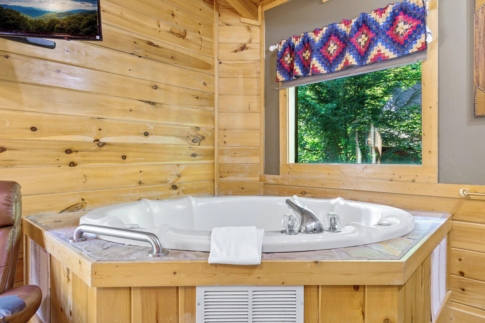A wooden cabin interior with a large bathtub, a window with a view of greenery, and a quilted curtain.
