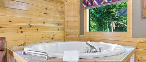 A wooden cabin interior with a large bathtub, a window with a view of greenery, and a quilted curtain.