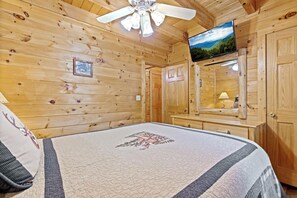 A wooden cabin room with a bed, a television, a ceiling fan, and a wooden dresser.