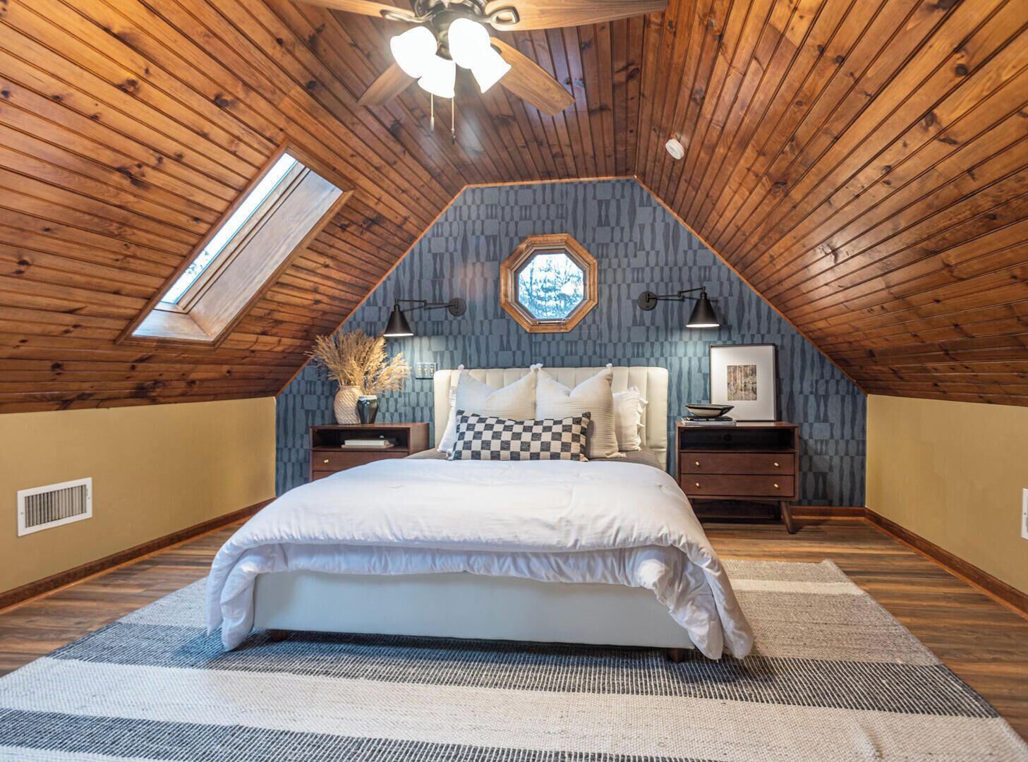 A bedroom with a sloped ceiling, a skylight, a large bed, two bedside tables, and a wall-mounted clock.