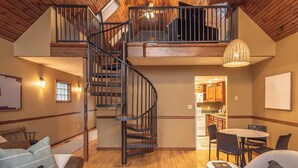 A spacious loft-style living area with a wooden ceiling, a spiral staircase, and a kitchen visible through an open doorway.