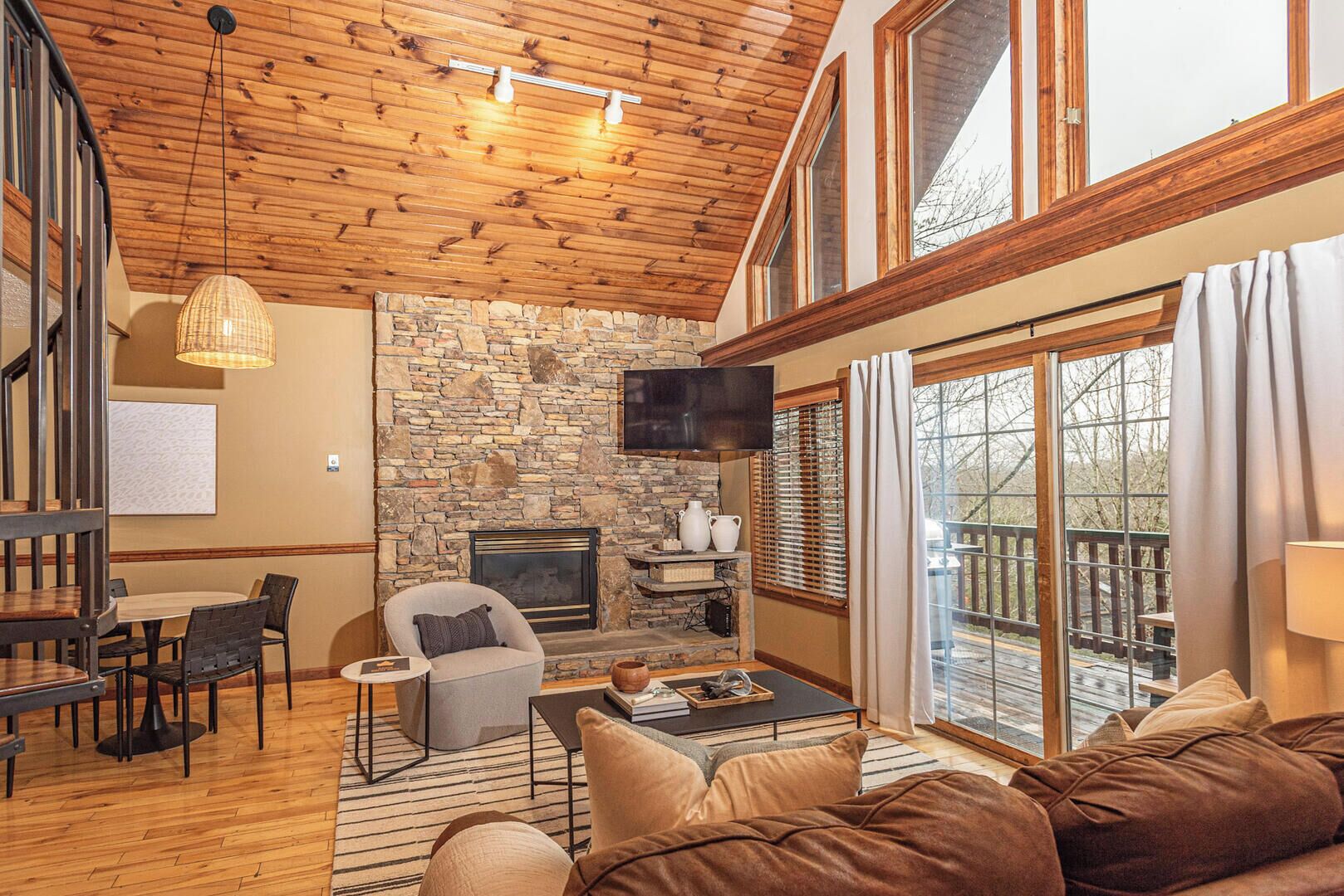 A cozy living room with a stone fireplace, wooden ceiling, and large windows.