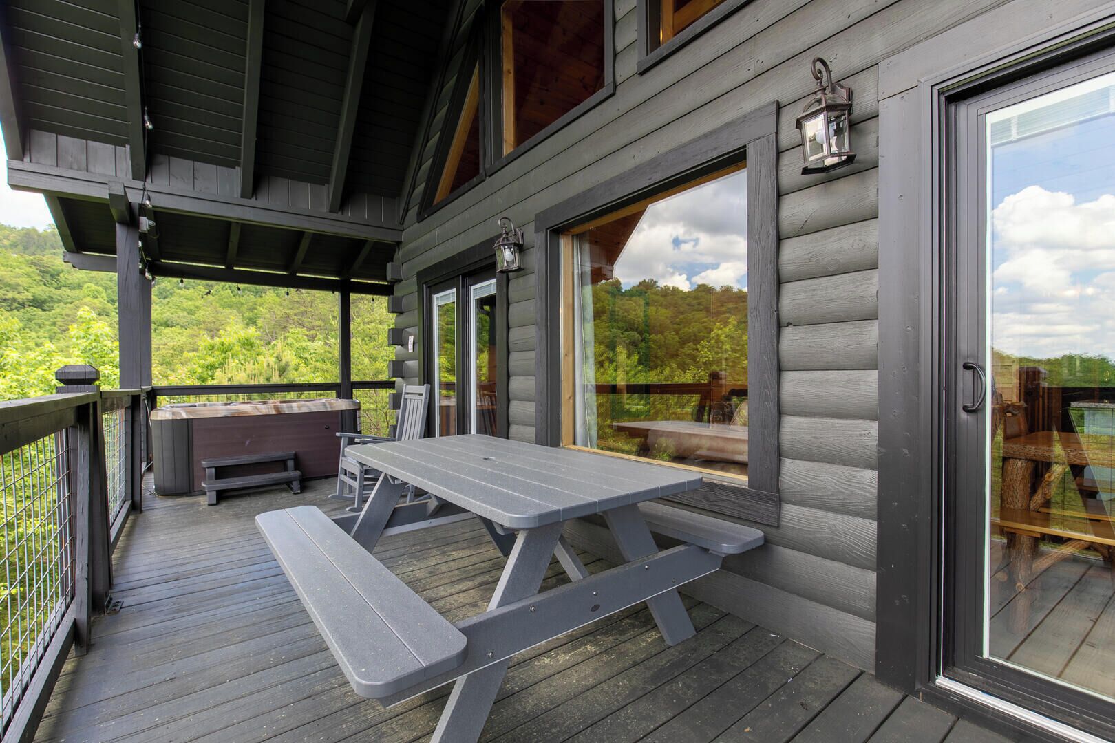 Une terrasse en bois avec une table de pique-nique et des bancs, un jacuzzi et une porte vitrée donnant sur une autre terrasse avec vue sur une zone boisée.