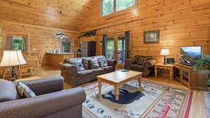 A spacious cabin with a high ceiling, wooden walls, a large window, a kitchen area, and a living room with a sofa, coffee table, and a television.