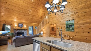 A cozy cabin interior with a stone fireplace, wooden walls, a dining area, and a kitchen with granite countertops.